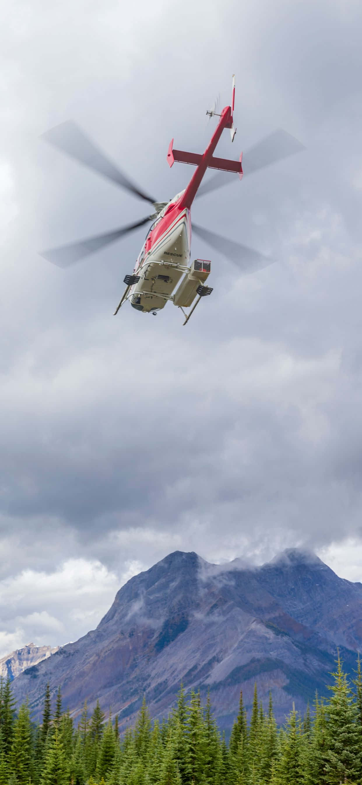 'n helikopter wat oor 'n bos vlieg met berge in die agtergrond Muurpapier