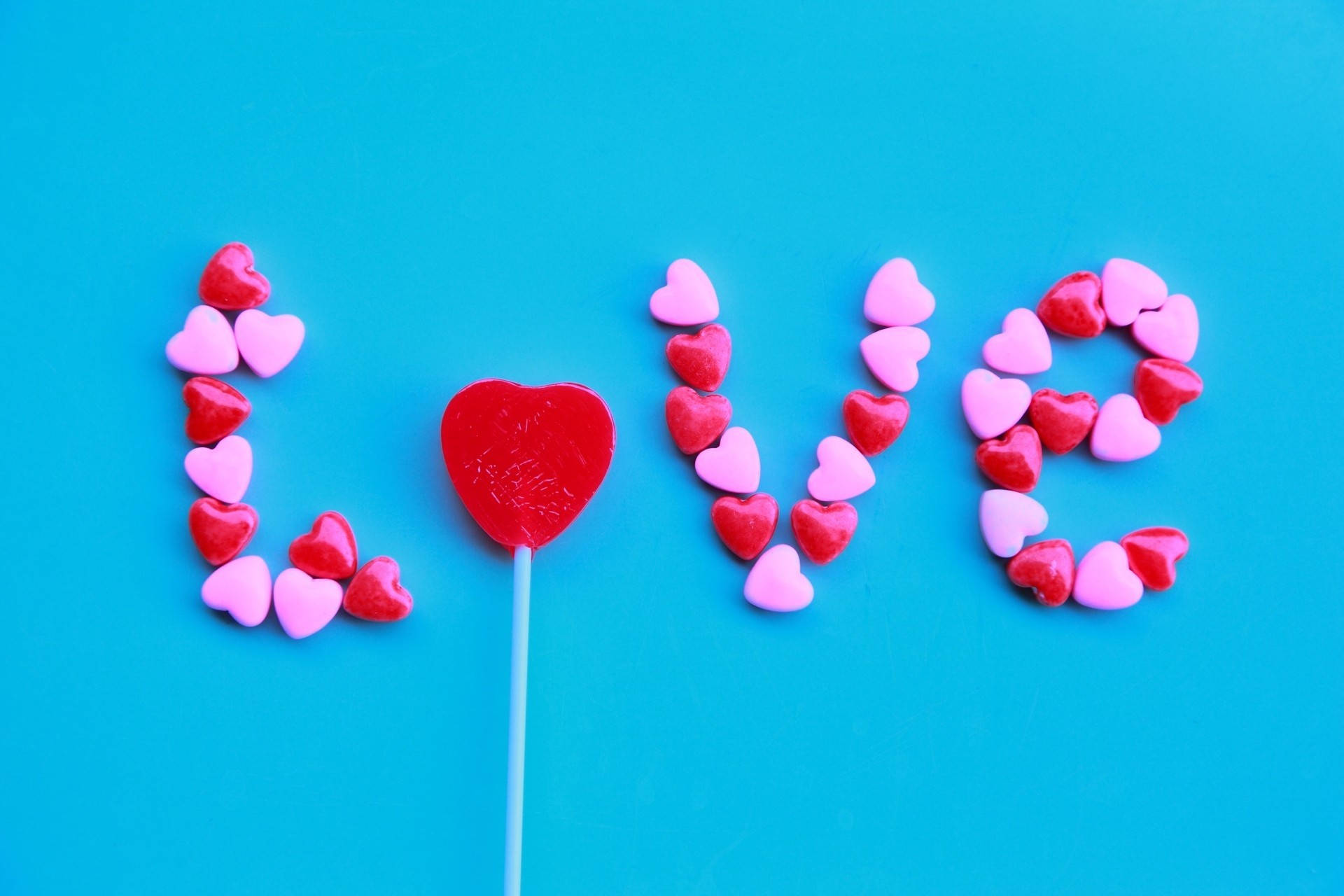 Pienk en rooi lekkergoedhartklippe wat die woord liefde skep op 'n helderblou muur vir Valentynsdag.