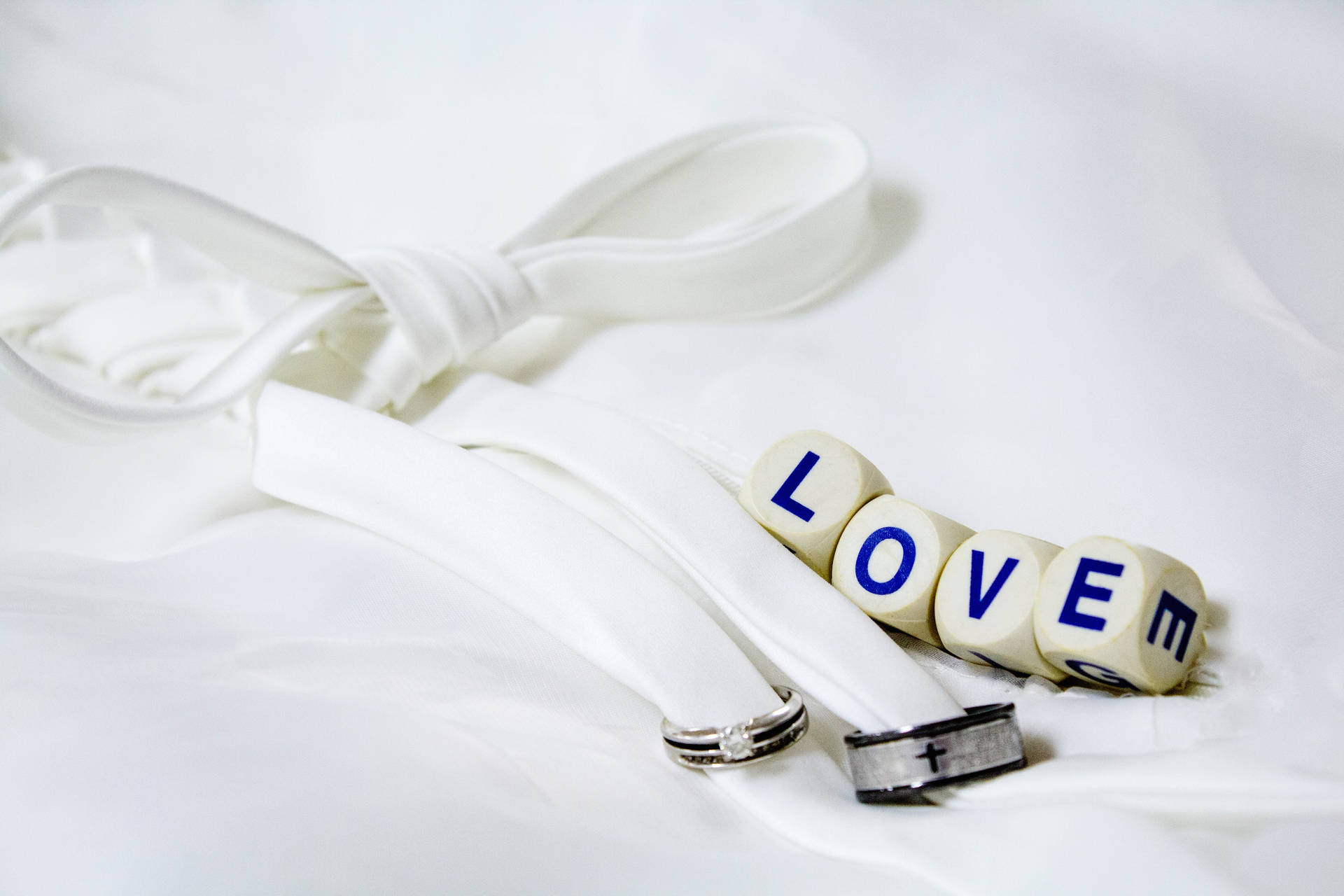 "Liefde" geskryf op vier wit dobbelstene langs twee silwer en swart trouringe wat met 'n wit lint vasgemaak is, geplaas op 'n wit laken.