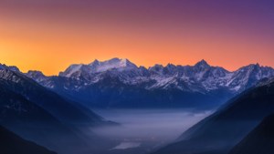 Asemrowende ultrawye uitsig oor sneeubedekte berge by sonsopkoms met mistige vallei.